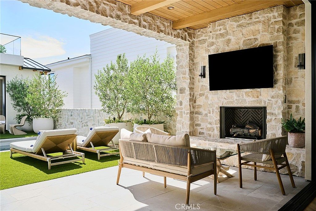 This covered outdoor living space features a stone-clad chimney breast with a built-in television and fireplace, creating a cozy focal point. The area is furnished with a light wood sofa, matching armchairs, and a glass-topped coffee table, all set on a paved patio overlooking a small, manicured lawn with potted olive trees. The space is open to the outdoors, blending indoor comfort with a bright, airy exterior atmosphere.