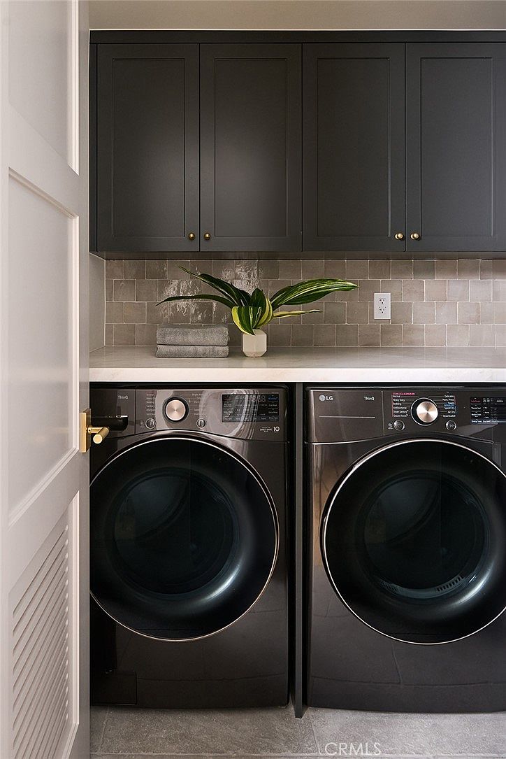 This modern laundry room features a pair of dark-toned front-loading appliances set beneath a clean, light-colored countertop. Above the machines, dark cabinetry provides ample storage, complemented by a neutral-toned tiled backsplash and a decorative plant. The space is framed by a white door on the left, creating a compact and functional utility area.