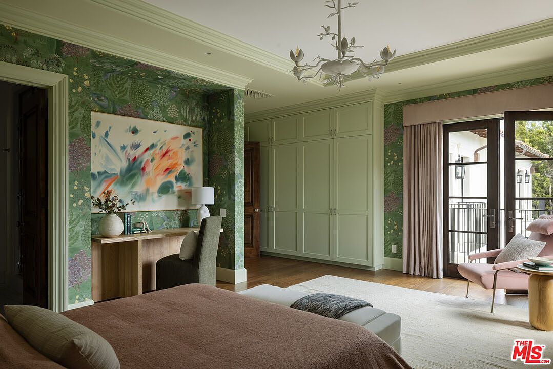 This elegant primary bedroom features a sophisticated blend of botanical-patterned wallpaper and soft green cabinetry, creating a serene and cohesive aesthetic. The room is furnished with a plush brown bed, a light wood desk area, and a soft pink armchair positioned near tall glass doors that open to a balcony. A delicate, light-colored chandelier hangs from the ceiling, adding a touch of refinement to the well-lit, spacious layout.