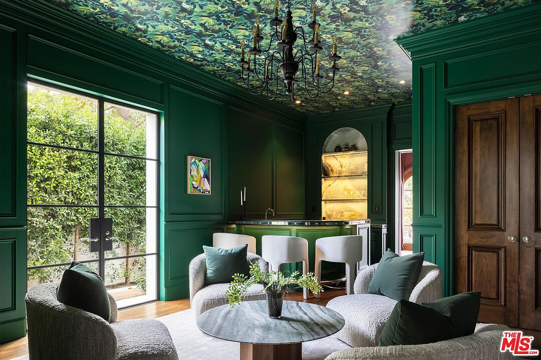 This sophisticated lounge area features deep green paneled walls and a striking botanical-patterned ceiling, creating a moody and luxurious atmosphere. The space is furnished with plush, textured armchairs arranged around a circular stone-topped coffee table, while a built-in bar with illuminated shelving serves as a focal point in the background. Large, multi-paned glass doors provide a view of the lush greenery outside, enhancing the room's elegant and intimate aesthetic.