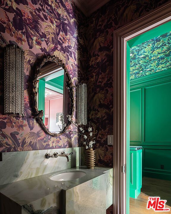 This powder room features bold, dark floral wallpaper and a stone-topped vanity with a wall-mounted faucet. An ornate, oval-framed mirror is flanked by two textured, vertical wall sconces, while a doorway leads into a vibrant green room with paneled walls and a matching ceiling treatment. The perspective captures the contrast between the moody, patterned foreground and the bright, saturated color palette of the adjacent space.