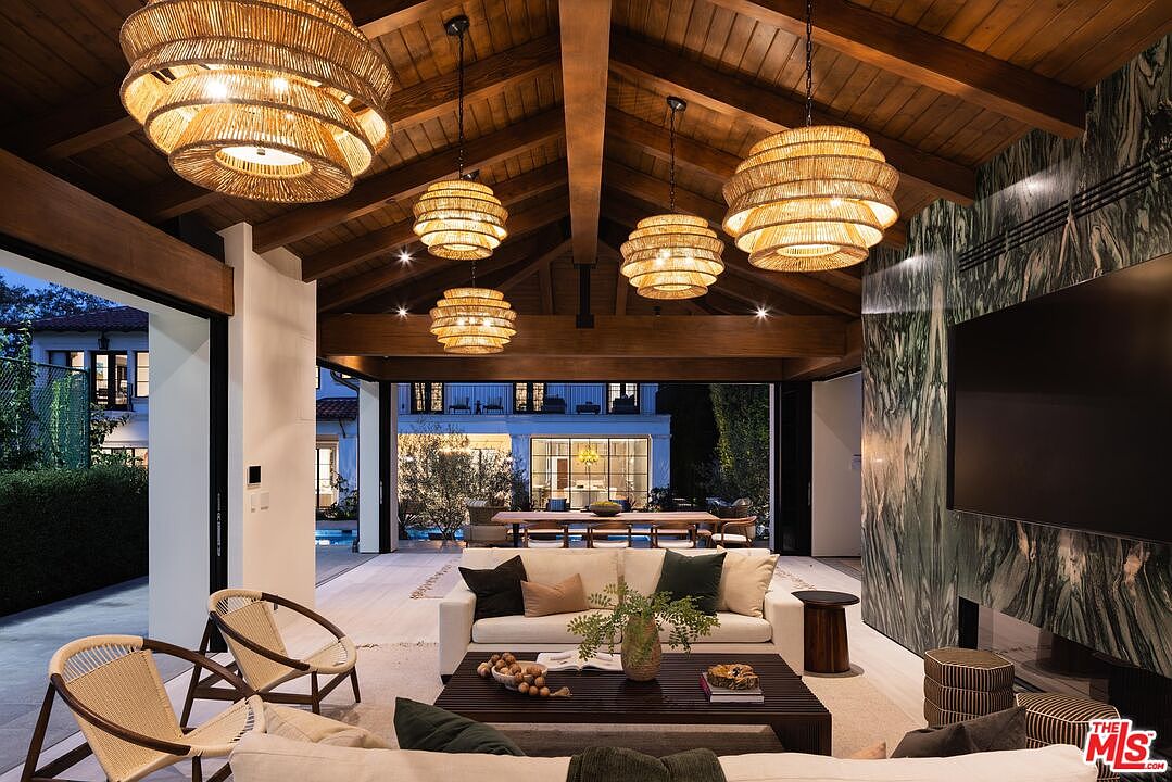 This expansive indoor-outdoor living space features a vaulted wood-paneled ceiling with exposed beams and multiple woven pendant lights. The room is anchored by a large sofa and a dark wood coffee table, set against a striking green stone-clad wall that houses a television. Large sliding glass doors open completely to reveal a dining area and a pool deck, creating a seamless transition between the interior and exterior environments.