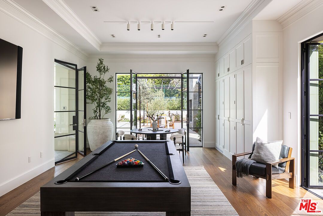 This sophisticated game room features a dark wood pool table as the central focal point, set upon a striped area rug. The space is brightened by white walls, extensive built-in cabinetry, and large glass-paned doors that open to an outdoor patio area. A modern armchair sits in the corner, and the room is illuminated by a sleek, linear track lighting fixture.