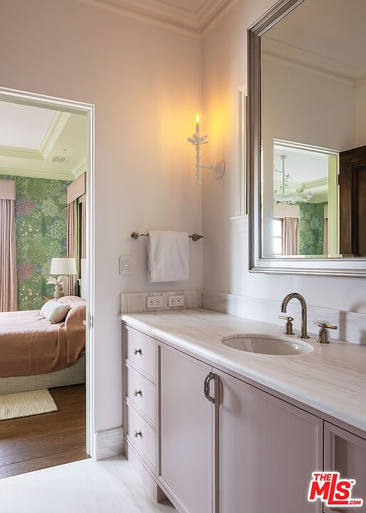 This elegant bathroom vanity area features a light-toned cabinet with a stone countertop and an integrated sink, complemented by a large, framed mirror. A wall-mounted candle-style sconce provides warm lighting, while a white towel hangs neatly on a rack nearby. The perspective looks through a doorway into an adjacent bedroom, which is decorated with floral wallpaper and soft pink curtains, creating a cohesive and sophisticated transition between spaces.