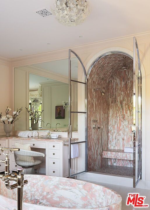 This luxurious primary bathroom features a striking arched shower enclosure with a glass door and intricate stone wall detailing. A matching stone-clad soaking tub sits in the foreground, complemented by a vanity area with a large mirror and elegant lighting. The space exudes a sophisticated, high-end aesthetic with a cohesive color palette and refined architectural elements.