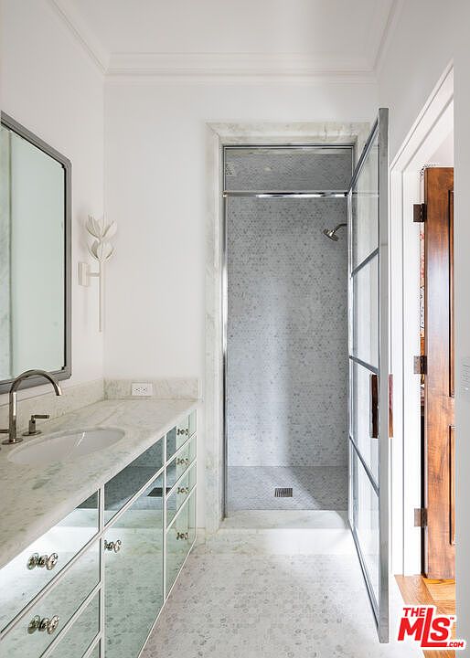This elegant bathroom features a vanity with mirrored cabinet doors and a light-toned stone countertop, complemented by a wall-mounted light fixture. A glass-enclosed shower with patterned tile walls and a pebble-style floor is positioned to the right, creating a bright and sophisticated atmosphere. The perspective looks directly into the shower area from the vanity, highlighting the clean lines and high-end finishes of the space.