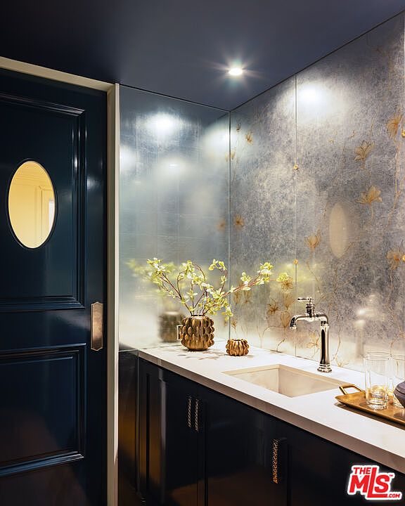 This sophisticated powder room features a dark, high-gloss navy door and cabinetry paired with a striking metallic-finish floral wallpaper backsplash. A sleek, light-toned stone countertop holds a modern faucet and a decorative vase, creating a luxurious and moody aesthetic. The perspective is a straight-on shot of the vanity area, highlighting the contrast between the dark finishes and the reflective wall treatment.
