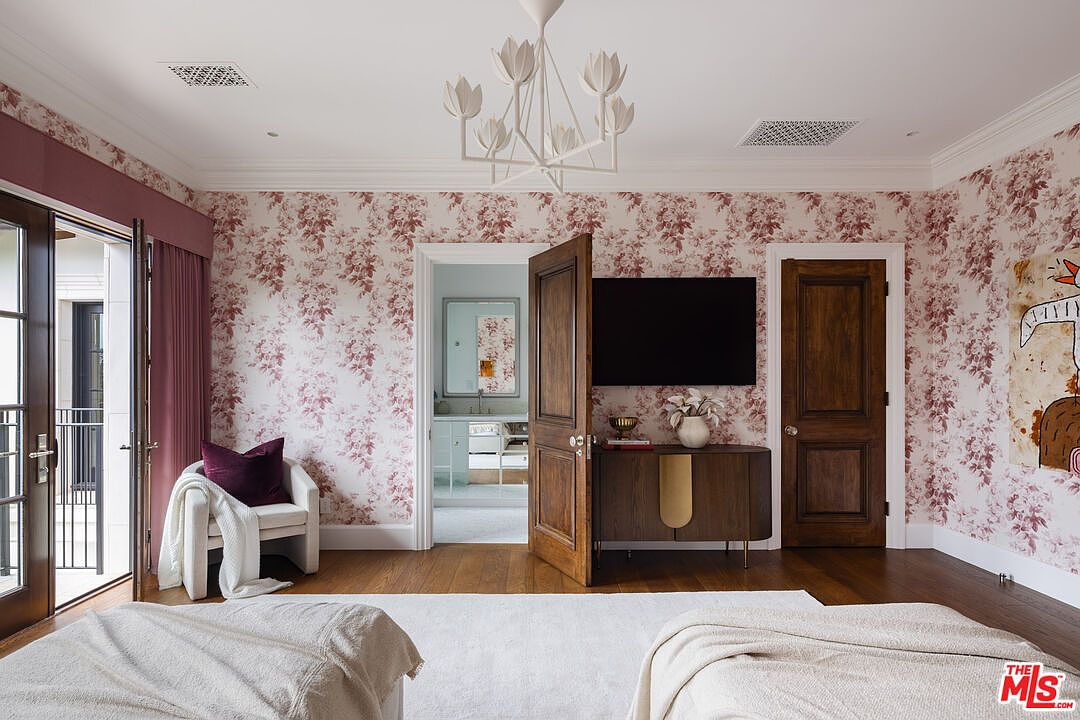 This elegant bedroom features floral wallpaper, a white chandelier, and a dark wood media console beneath a wall-mounted television. A doorway leads into an adjacent bathroom, while a glass door on the left opens to a balcony, allowing natural light to fill the space. The room is furnished with a comfortable armchair and plush bedding, creating a sophisticated and inviting atmosphere.