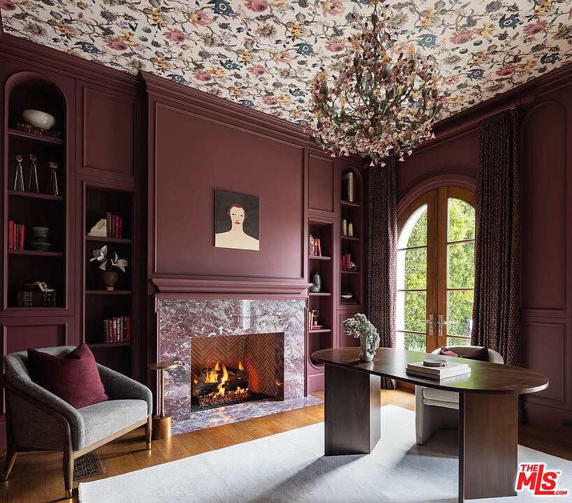 This sophisticated home office features deep plum-colored paneled walls and a striking floral wallpaper on the ceiling. A central fireplace with a stone surround serves as the focal point, flanked by built-in shelving units, while a modern desk and a gray armchair provide a functional workspace. The room is bathed in natural light from arched glass doors that lead to the outdoors, creating an elegant and moody atmosphere.