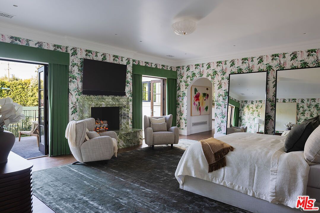 This spacious primary bedroom features a sophisticated design with bold floral wallpaper and a large, dark area rug anchoring the seating area. A fireplace with a striking green stone surround serves as a focal point, flanked by two comfortable armchairs, while two large mirrors lean against the wall near the bed. The room opens onto a private balcony through glass doors, creating a seamless transition between the elegant interior and the outdoor space.