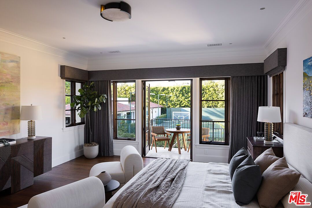 This elegant primary bedroom features a neutral color palette with a plush bed, a modern curved lounge chair, and dark wood flooring. Large glass doors open onto a private balcony that overlooks a tennis court, seamlessly blending indoor and outdoor living. The room is accented by sophisticated window treatments, matching bedside lamps, and a sleek console table, creating a serene and luxurious atmosphere.