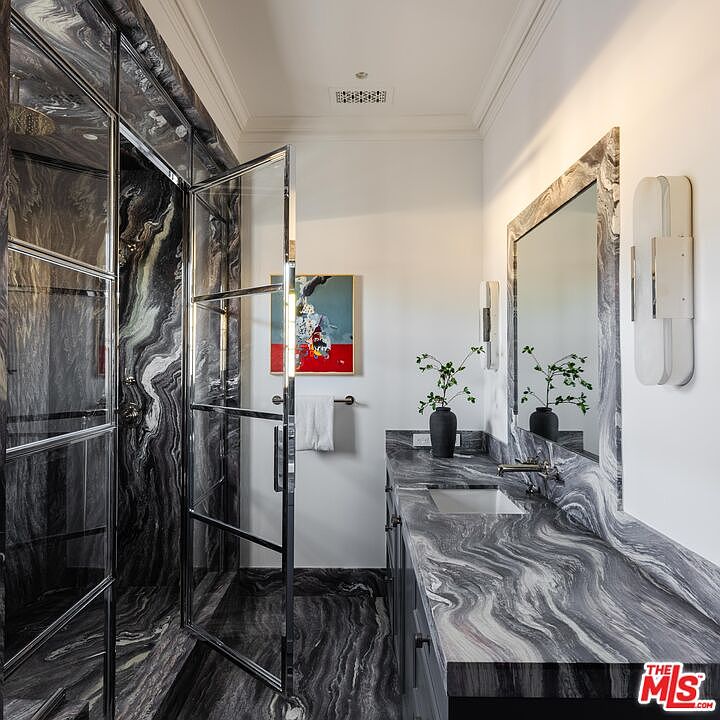 This sophisticated bathroom features striking, high-contrast patterned stone surfaces on the vanity countertop and shower walls, creating a dramatic and luxurious aesthetic. A glass-enclosed shower with a sleek metal frame stands adjacent to a vanity with a mirror and modern wall-mounted lighting. The space is accented by a vibrant piece of wall art and a small potted plant, offering a refined and contemporary atmosphere.