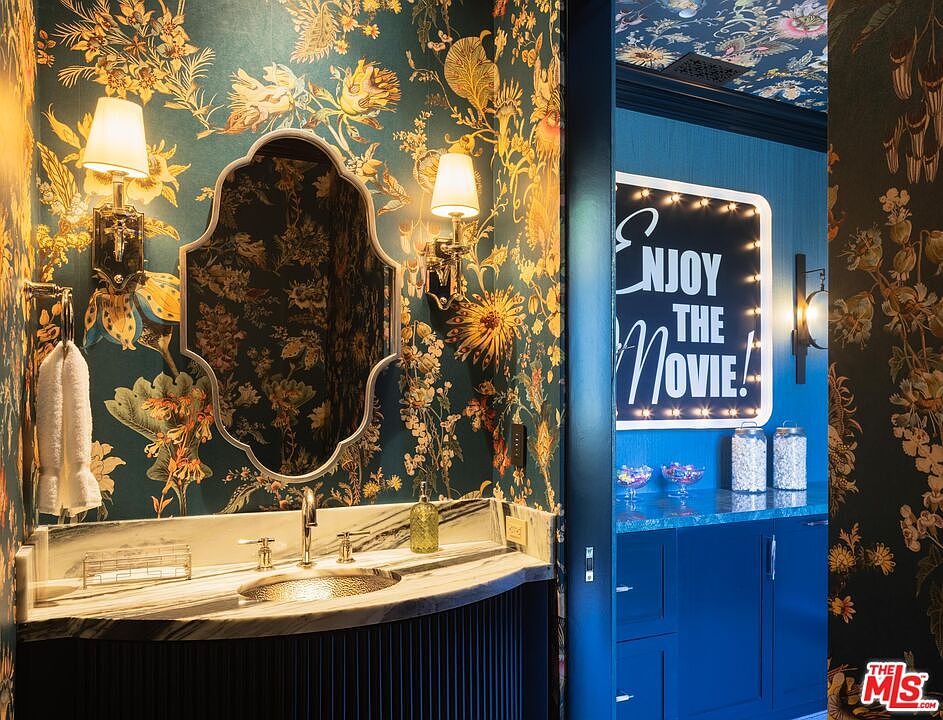 This vibrant powder room features bold, dark floral wallpaper and a uniquely shaped mirror flanked by elegant wall sconces. The vanity is topped with a polished stone surface and a metallic basin, while an adjacent doorway reveals a playful, illuminated 'Enjoy the Movie' sign in a blue-toned media or snack area. The perspective captures the transition between the ornate bathroom and the adjacent entertainment space, highlighting a sophisticated and eclectic design aesthetic.