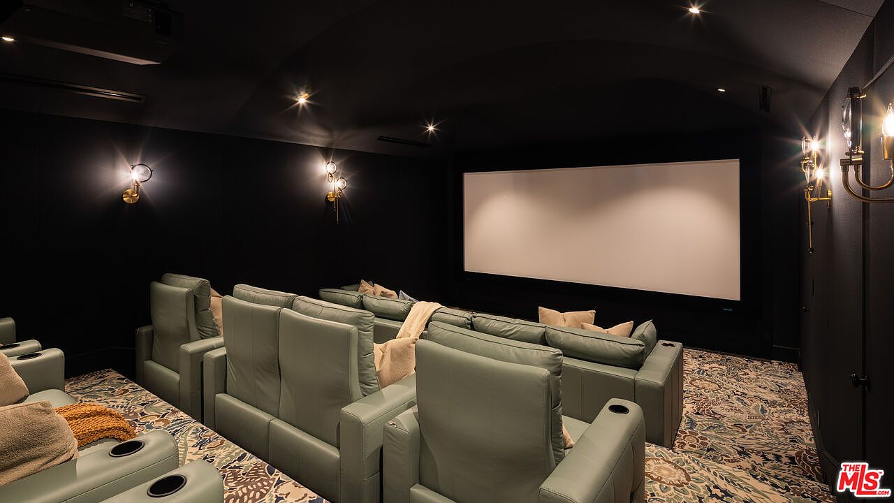 This dedicated home theater room features tiered rows of plush, light-colored leather seating arranged for optimal viewing of a large projection screen. The space is designed with dark, moody walls and a vaulted ceiling, complemented by elegant wall-mounted sconces that provide soft, ambient lighting. A patterned carpet covers the floor, adding texture to the sophisticated and immersive cinematic environment.