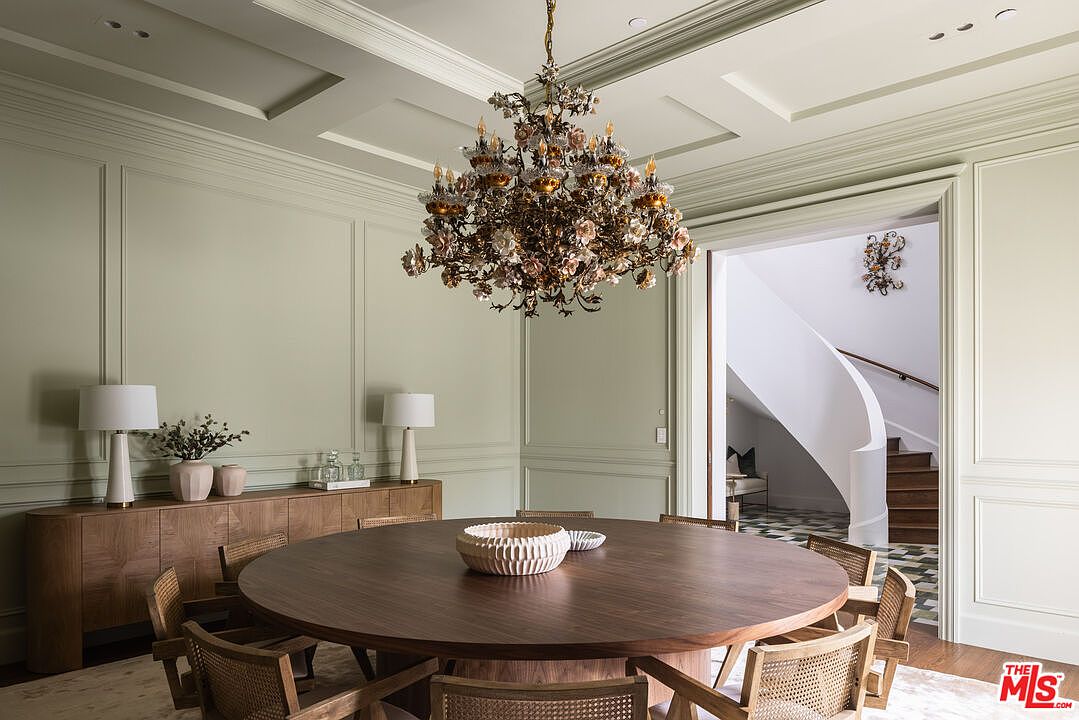 This elegant dining room features a large, round wooden table centered beneath an ornate, multi-tiered floral chandelier. The walls are adorned with classic wainscoting and painted in a soft sage green, while a long wooden sideboard provides storage and display space. A wide doorway leads to a bright hallway with a curved staircase, creating a sophisticated and open transition between spaces.