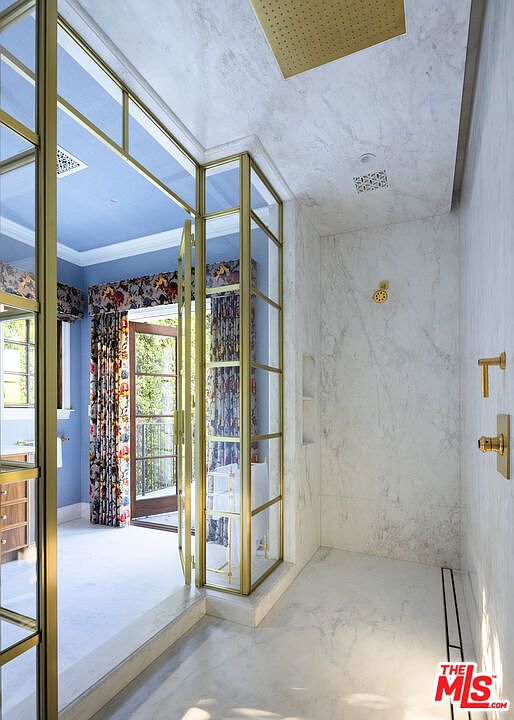 This luxurious primary bathroom features a walk-in shower enclosed by a sophisticated brass-framed glass partition. The shower area is finished with light-toned stone surfaces and includes a rainfall showerhead, while the adjacent room showcases vibrant floral curtains and a blue ceiling. The perspective looks from the shower outward, highlighting the elegant transition between the spa-like bathing space and the adjoining bedroom or dressing area.
