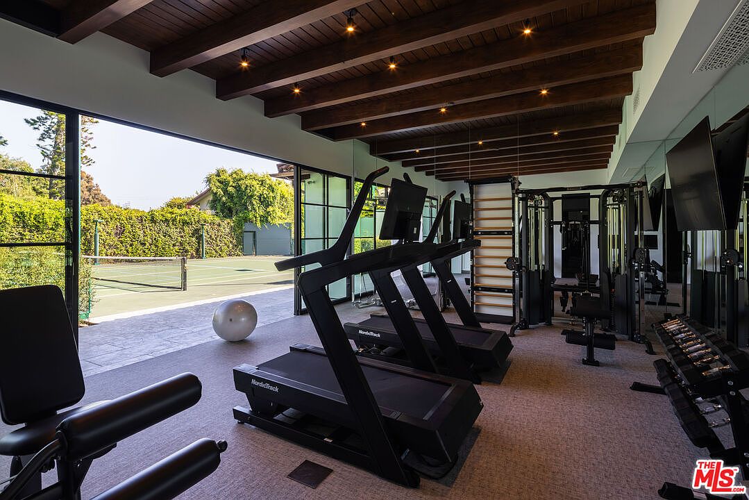 This spacious home gym features a high-end, modern aesthetic with exposed dark wood ceiling beams and recessed lighting. The room is equipped with multiple treadmills and a weight training station, while large glass doors open directly onto an outdoor tennis court. The combination of professional-grade fitness equipment and the seamless transition to the exterior creates an inviting and functional workout environment.