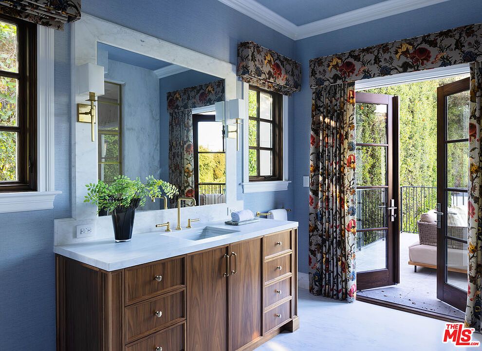 This elegant primary bathroom features a wood vanity with a light-colored stone countertop and a large, framed mirror. The room is painted in a soft blue, complemented by floral-patterned window treatments and curtains that frame a set of French doors leading to an outdoor balcony. The space combines classic design elements with a bright, airy atmosphere, offering a seamless transition between the indoor vanity area and the exterior living space.