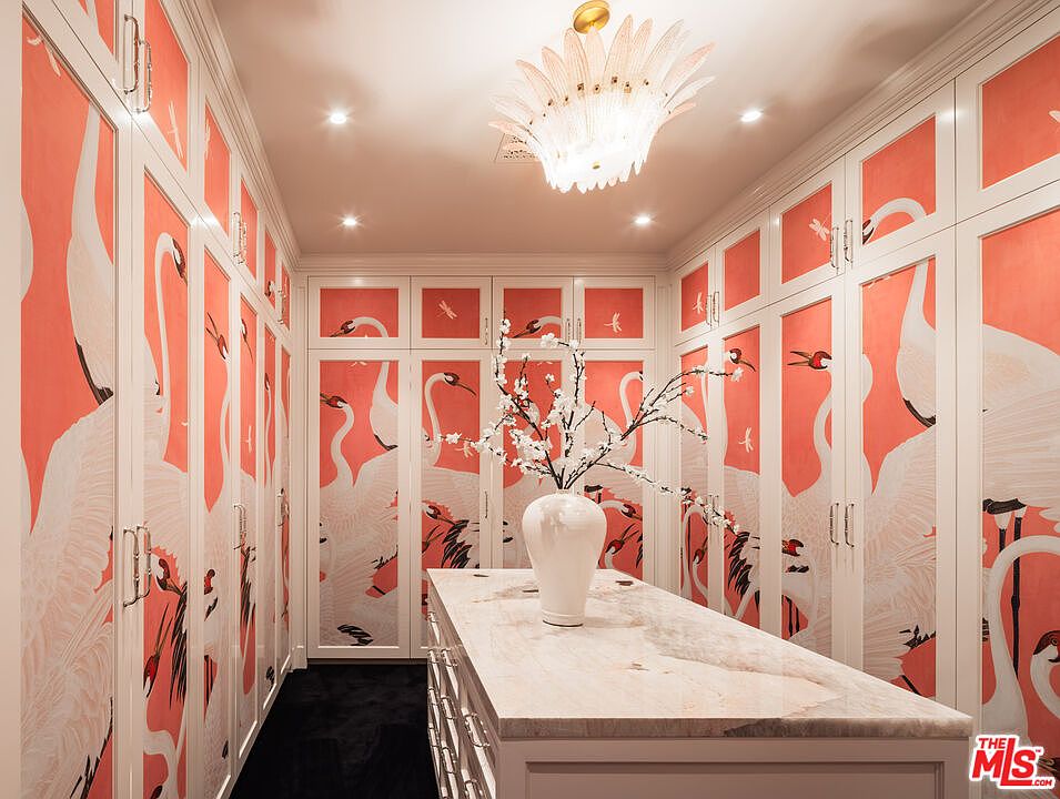 This luxurious walk-in closet features custom cabinetry adorned with a vibrant, artistic crane-patterned wallpaper. A central island with a stone countertop provides a focal point, topped with a decorative vase of white blossoms, while a unique, feathered-style chandelier hangs from the ceiling. The space is framed by dark flooring and symmetrical storage units, creating a sophisticated and highly stylized dressing area.