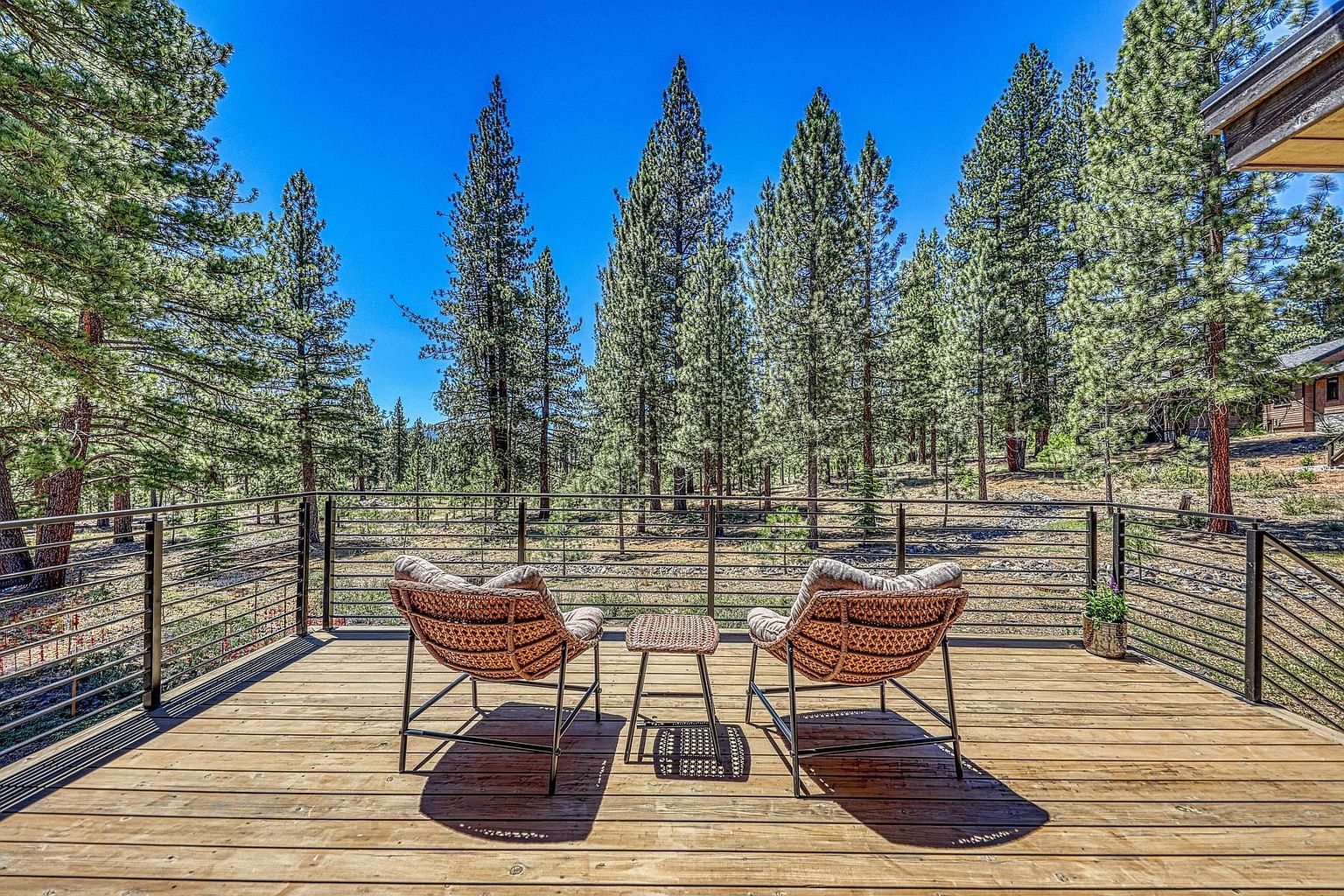 This elevated wooden deck offers a serene outdoor retreat, featuring two woven armchairs and a small side table positioned to overlook a dense forest of pine trees. The deck is enclosed by a modern metal railing, providing an unobstructed view of the natural landscape under a clear blue sky. The perspective is eye-level, capturing the inviting atmosphere of a peaceful mountain or woodland getaway.