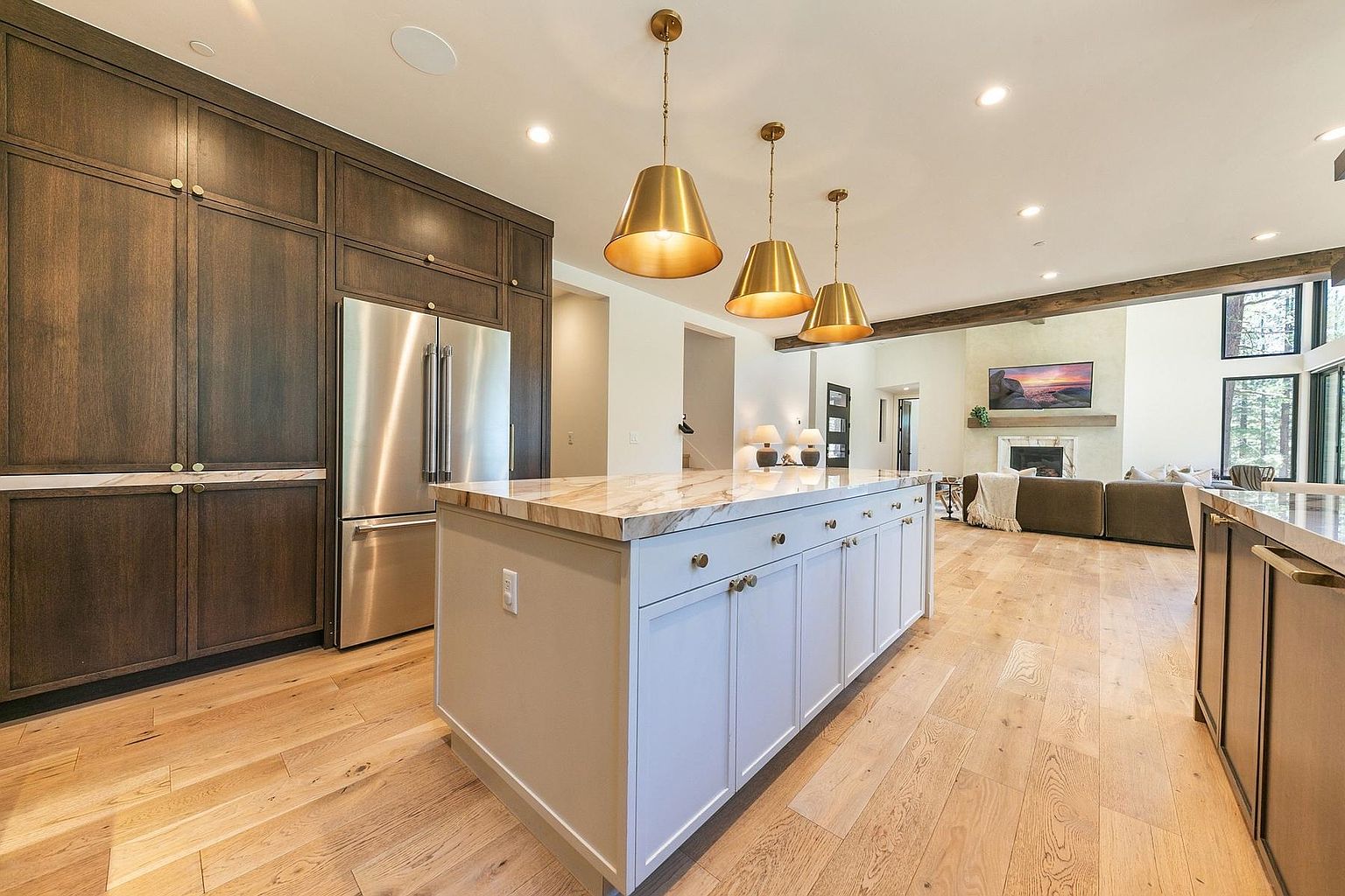 This open-concept kitchen features a large central island with a light-toned stone countertop and a light gray base, illuminated by three metallic pendant lights. Dark wood cabinetry provides a striking contrast against the light wood flooring, while the space flows seamlessly into a bright living area with a fireplace. The perspective captures the transition between the functional kitchen workspace and the comfortable, airy living room beyond.