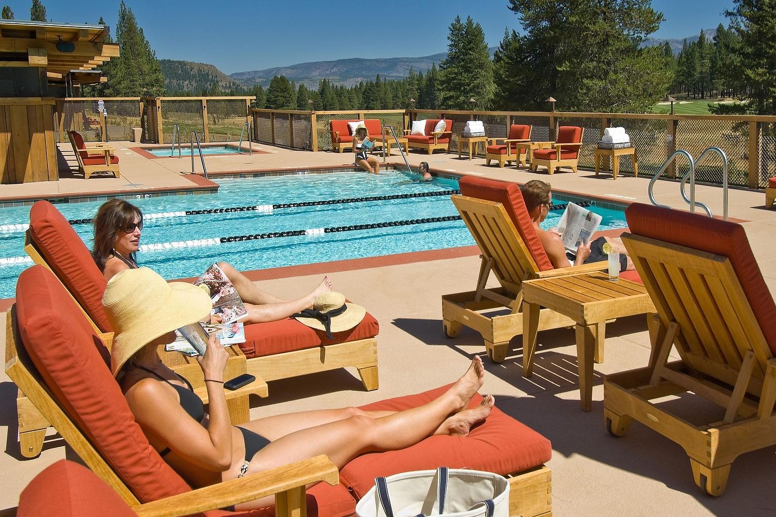 This inviting outdoor pool area features a lap pool surrounded by a spacious deck furnished with comfortable wooden lounge chairs and plush cushions. The scene is set against a backdrop of lush pine trees and distant mountains, creating a serene and resort-like atmosphere. Several people are relaxing by the water, emphasizing the leisure and recreational appeal of this community amenity.