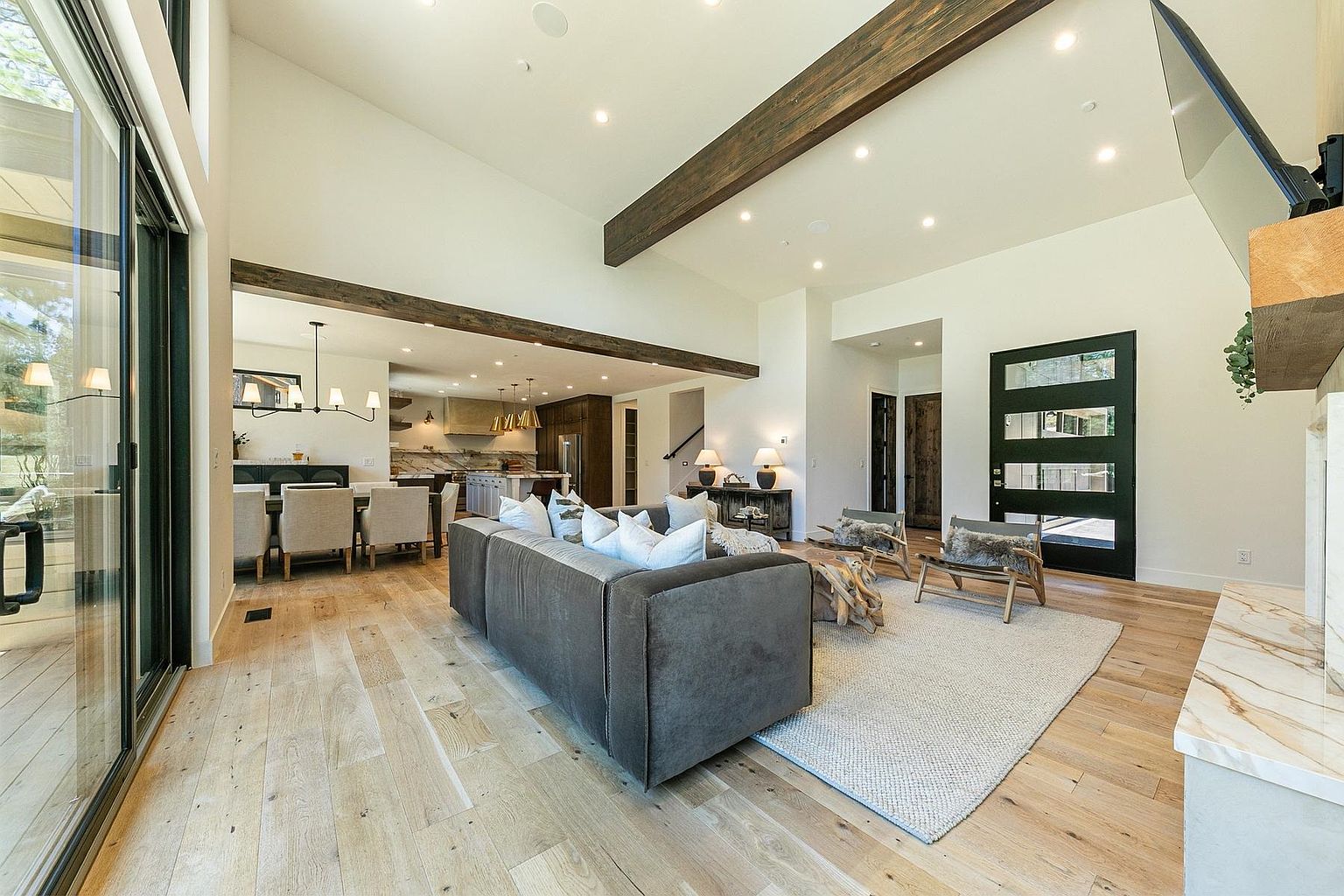 This open-concept living area features light wood flooring, exposed dark wood ceiling beams, and a neutral color palette that creates a bright, airy atmosphere. The space includes a large sectional sofa, two accent chairs, and a central coffee table, all arranged on a textured area rug. A tall stone-clad fireplace surround is visible on the right, while the background reveals a dining area and kitchen, emphasizing the home's spacious and cohesive design.