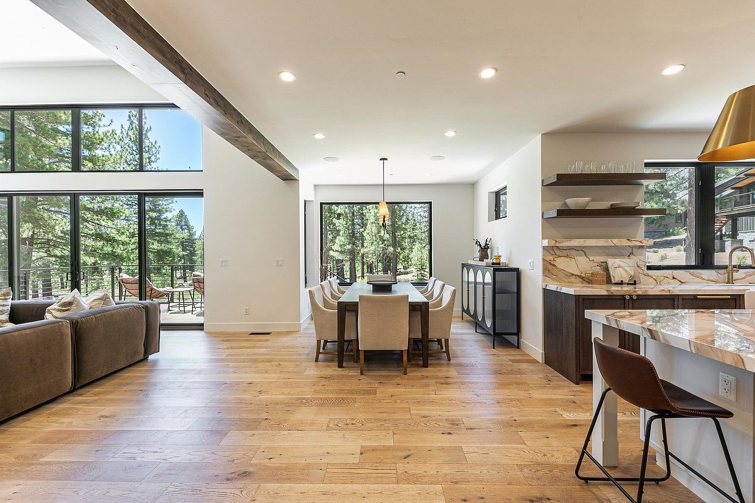 This open-concept living space features a dining area as the central focal point, seamlessly connecting to a kitchen on the right and a living area on the left. The room is anchored by a long dining table surrounded by upholstered chairs, illuminated by a minimalist pendant light, with light wood flooring extending throughout the entire area. Large windows and glass doors provide an expansive view of the surrounding forest, creating a bright, airy atmosphere with a modern, nature-inspired aesthetic.