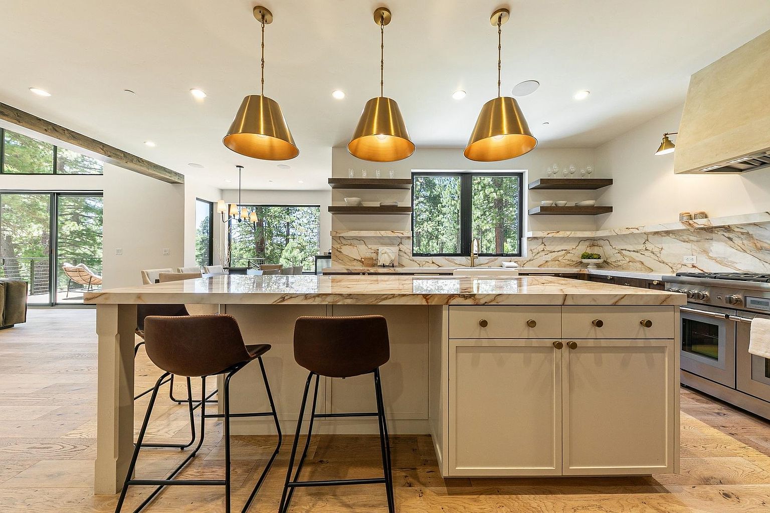 This modern kitchen features a large central island with a stone countertop and two leather-upholstered bar stools. Three metallic pendant lights hang above the island, which faces a windowed wall with floating wood shelves and a stone backsplash. The space is bright and open, showcasing light wood flooring and a professional-grade stainless steel range, creating a sophisticated and airy atmosphere.