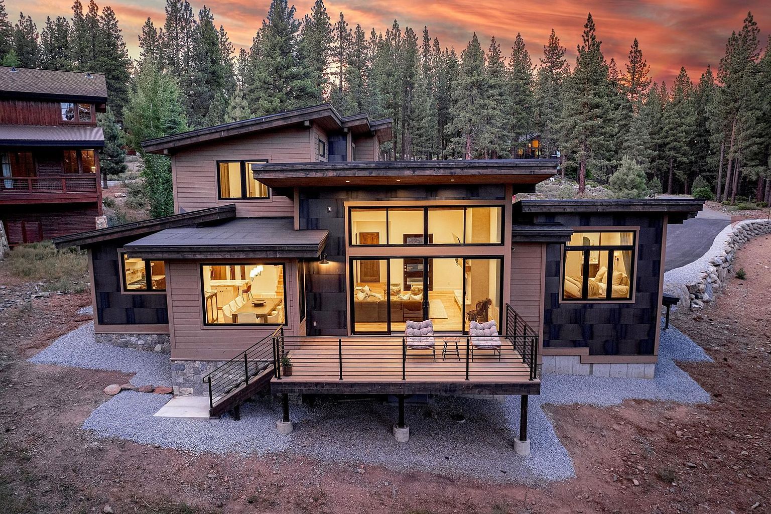 This modern mountain home features a striking blend of horizontal wood siding and dark, textured paneling, set against a backdrop of dense pine trees at sunset. The rear elevation showcases a spacious wooden deck with metal railings, large windows, and sliding glass doors that provide a seamless connection between the indoor living areas and the natural surroundings. The architectural design emphasizes clean lines and a contemporary aesthetic, perfectly integrated into the rugged, forested landscape.