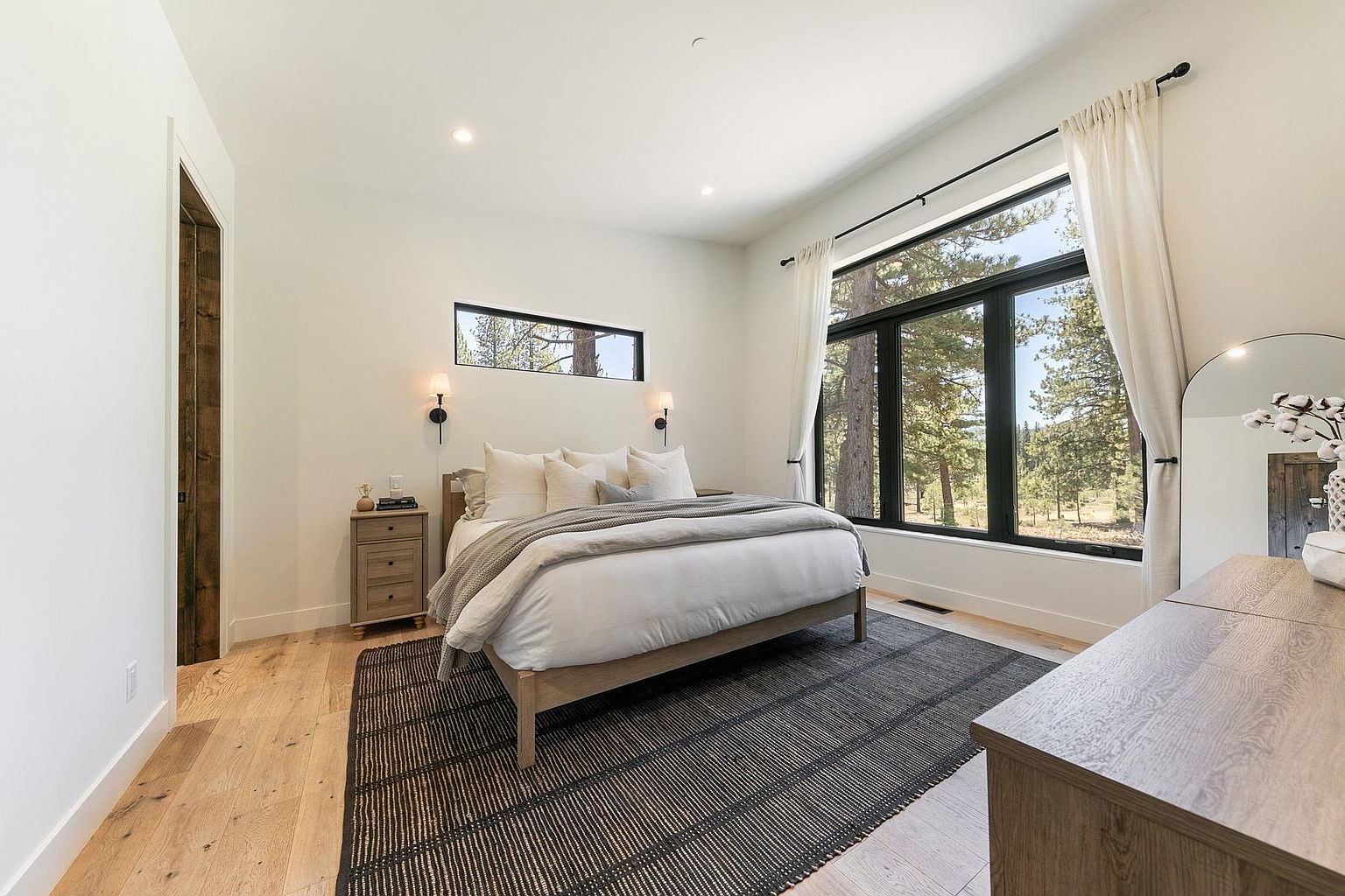 This bright and airy primary bedroom features light wood flooring and a neutral color palette, centered around a bed dressed in white linens and a textured throw. A large window with black framing provides a scenic view of the surrounding forest, complemented by white curtains and a horizontal accent window above the headboard. The room is furnished with a matching light wood nightstand and a dresser with an arched mirror, creating a cohesive and serene atmosphere.
