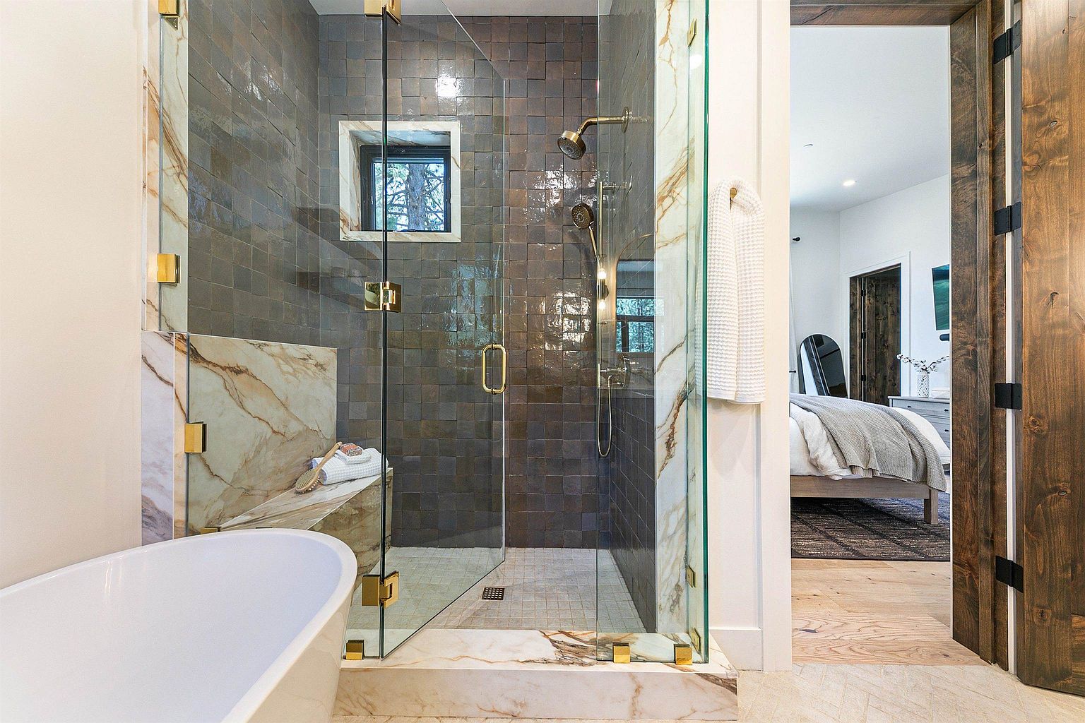This luxurious bathroom features a glass-enclosed walk-in shower with dark, textured square tiles and a built-in stone bench. A freestanding white soaking tub sits in the foreground, while a doorway provides a clear line of sight into an adjacent bedroom with a neutral color palette. The space is accented with gold-toned hardware and fixtures, creating a sophisticated and high-end aesthetic.