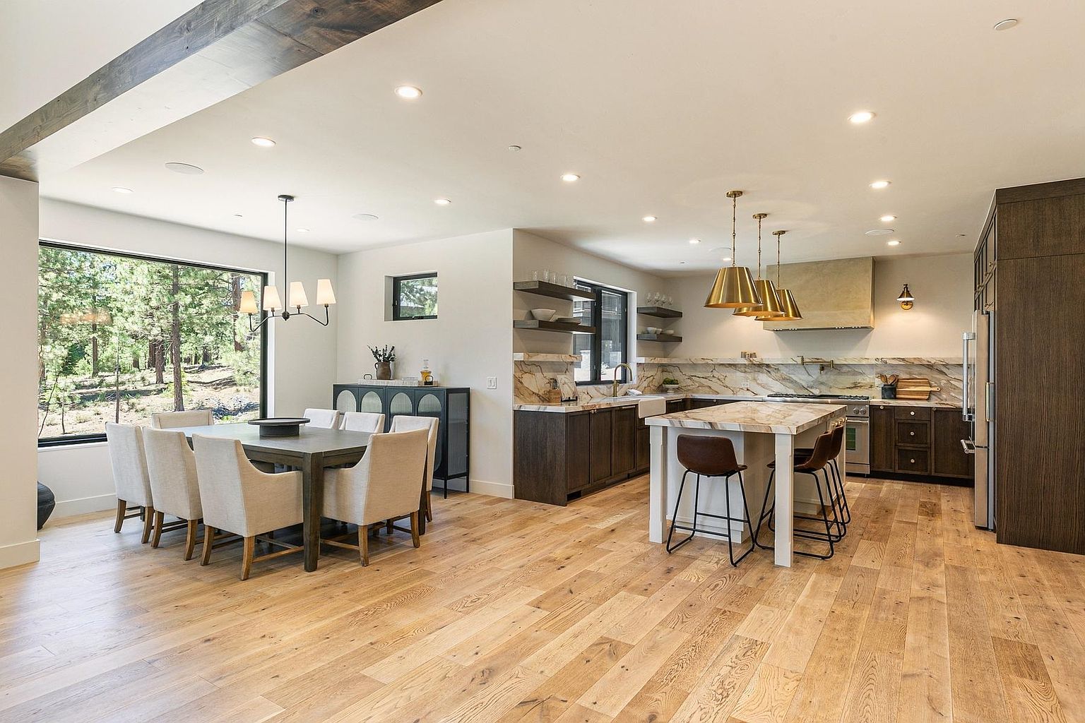 This open-concept space features a modern kitchen and dining area with light wood flooring and a neutral color palette. The kitchen is equipped with dark cabinetry, a large island with bar seating, and stone countertops that extend to the backsplash. A dining table with upholstered chairs sits near a large window, creating a bright and inviting atmosphere that flows seamlessly into the culinary workspace.