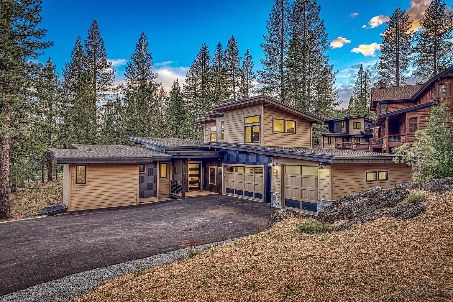 This modern mountain home features a multi-level design with horizontal wood siding and dark trim, nestled among tall pine trees. The front facade includes a covered entryway, a two-car garage with glass-paneled doors, and a paved driveway leading up to the house. The exterior is accented by natural stone elements and large windows, creating a sophisticated, contemporary aesthetic in a serene, wooded setting.
