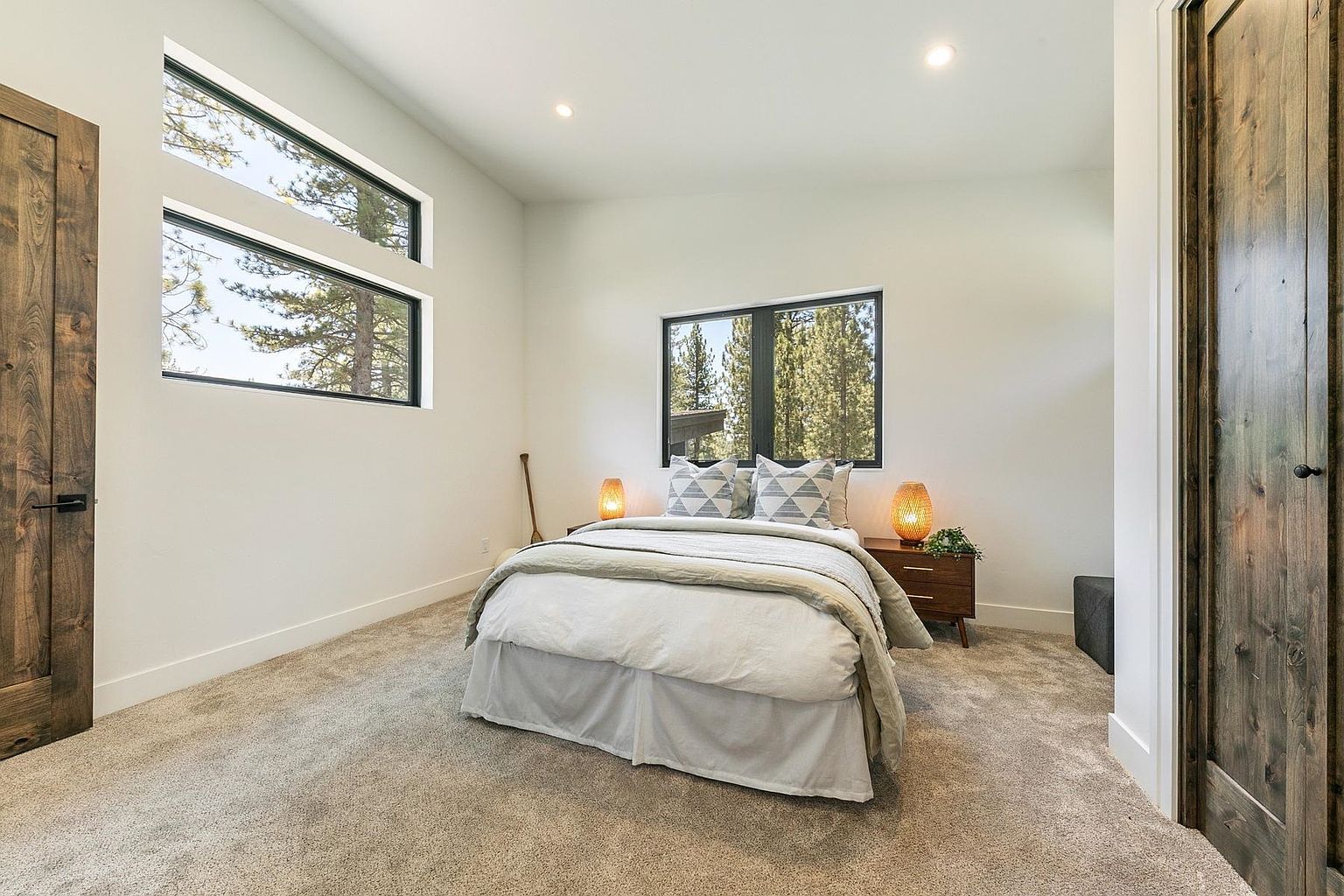 This bright and airy bedroom features neutral carpeting and crisp white walls, creating a clean, modern aesthetic. The room is centered around a bed with light-toned bedding and patterned pillows, flanked by two wooden nightstands topped with warm, glowing lamps. Large windows on two walls provide ample natural light and views of the surrounding trees, while rustic wood-toned doors add a touch of warmth to the space.