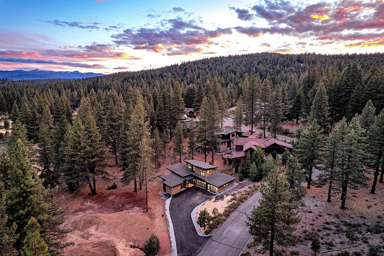 This high-angle aerial perspective captures a modern mountain residence nestled within a dense pine forest at sunset. The home features a contemporary design with multi-level flat rooflines, large windows, and a paved driveway that curves through the wooded landscape. The dramatic sky, filled with vibrant pink and orange clouds, creates a serene and picturesque backdrop for this secluded property.