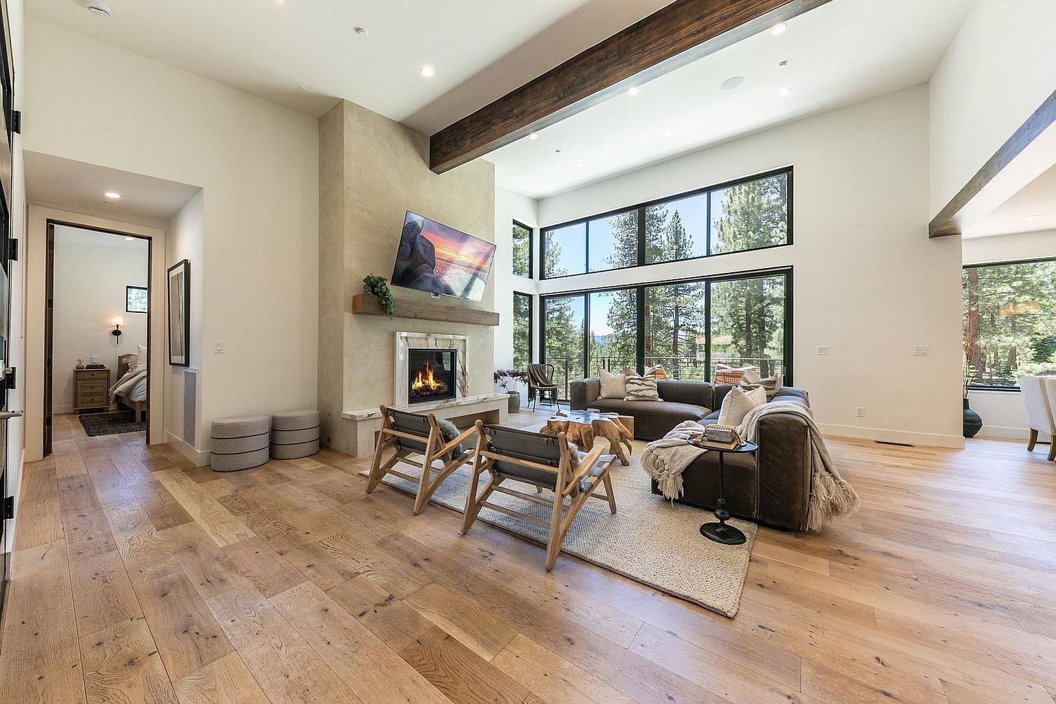 This spacious living room features light wood flooring, a large sectional sofa, and two wooden armchairs arranged around a unique natural wood coffee table. A stone-clad chimney breast serves as the focal point, housing a fireplace with a wooden mantel and a television mounted above. Large windows provide an expansive view of the surrounding forest, while a dark wooden ceiling beam adds architectural interest to the bright, open-concept space.
