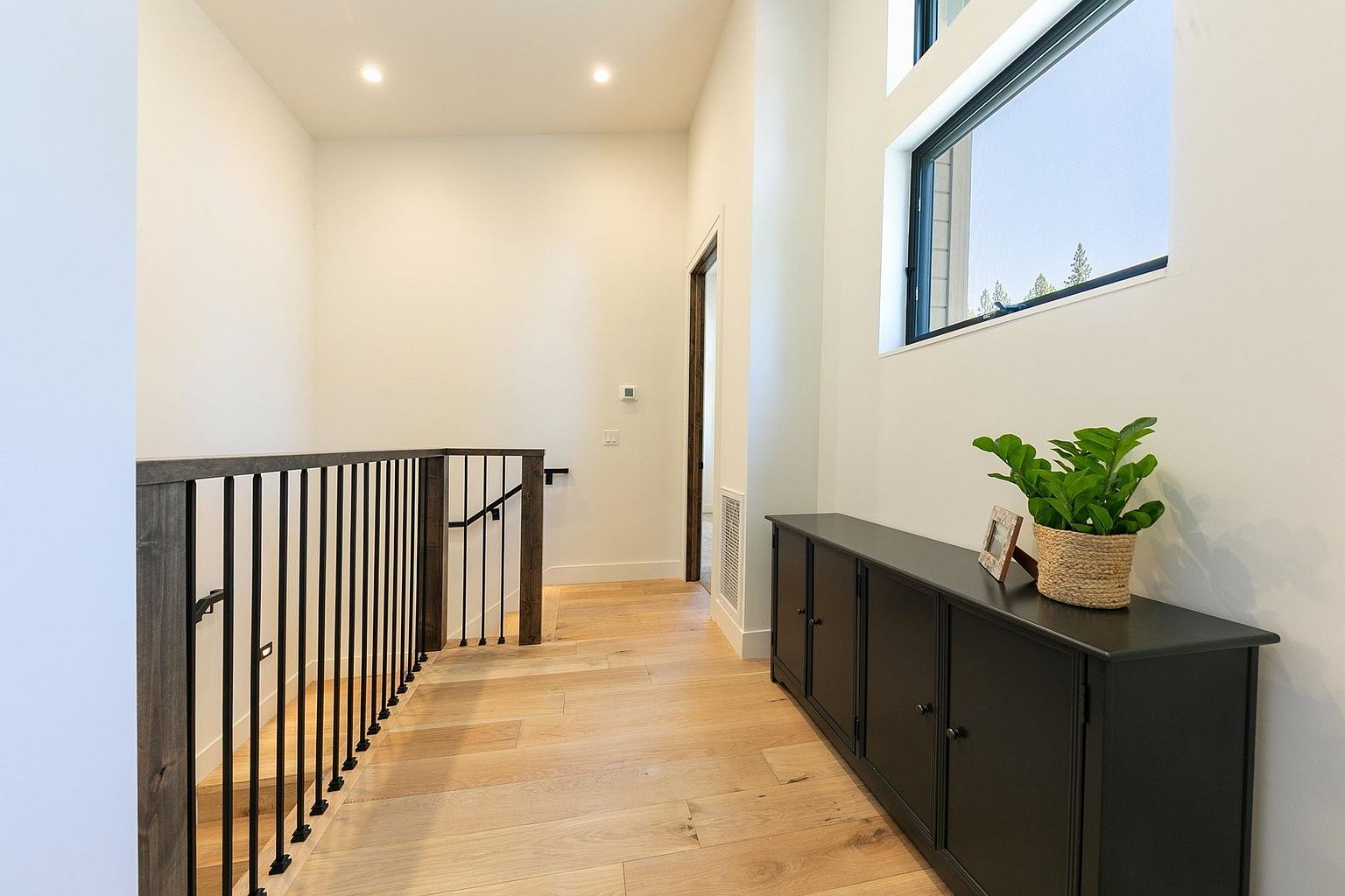 This bright, modern hallway features light wood flooring and a sleek black metal railing overlooking the staircase. A long, black console table sits against the wall, topped with a small potted plant and a picture frame, while a large window allows natural light to fill the space. The clean lines and neutral color palette create an airy, contemporary atmosphere.