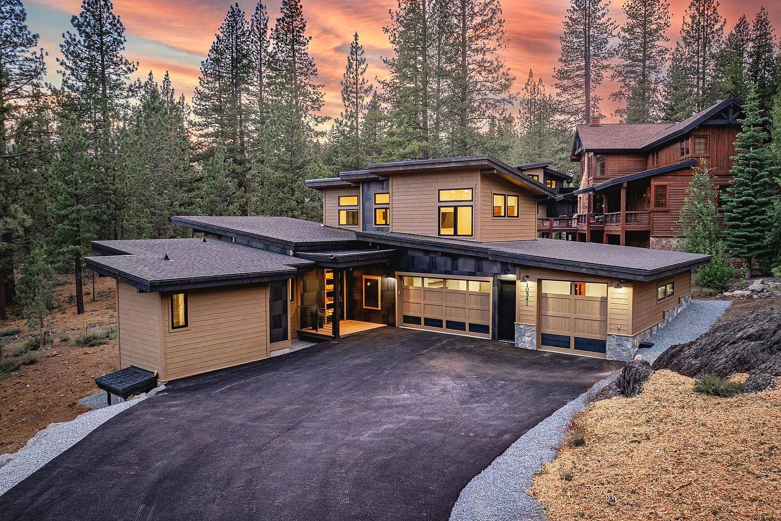 This modern mountain home features a striking architectural design with clean lines, a multi-level shed roof, and a blend of horizontal wood siding and stone accents. The front of the property includes a spacious paved driveway leading to a two-car garage, with a welcoming entryway tucked under a covered porch. Set against a backdrop of dense pine trees at sunset, the home exudes a sophisticated, contemporary aesthetic that blends seamlessly with its natural surroundings.