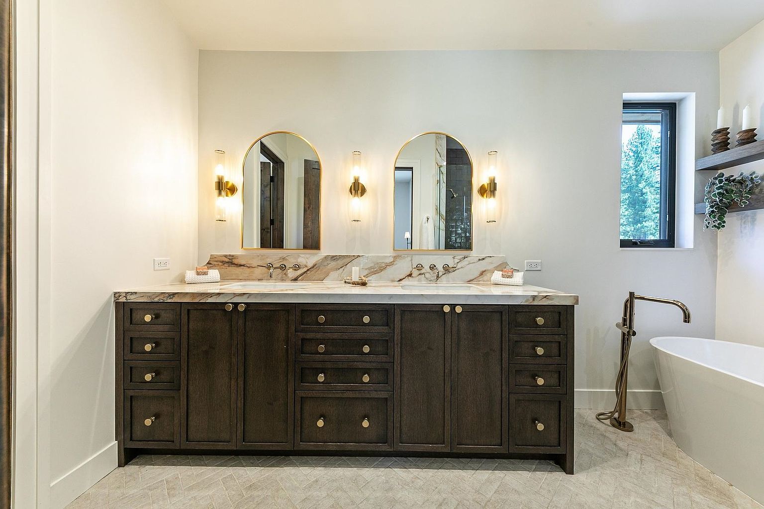 This elegant primary bathroom features a double vanity with dark wood cabinetry and a light-toned stone countertop with a matching backsplash. Two arched mirrors are mounted above the vanity, flanked by modern gold-toned wall sconces, while a freestanding soaking tub sits to the right near a window. The space is finished with light-colored herringbone patterned flooring, creating a sophisticated and cohesive aesthetic.