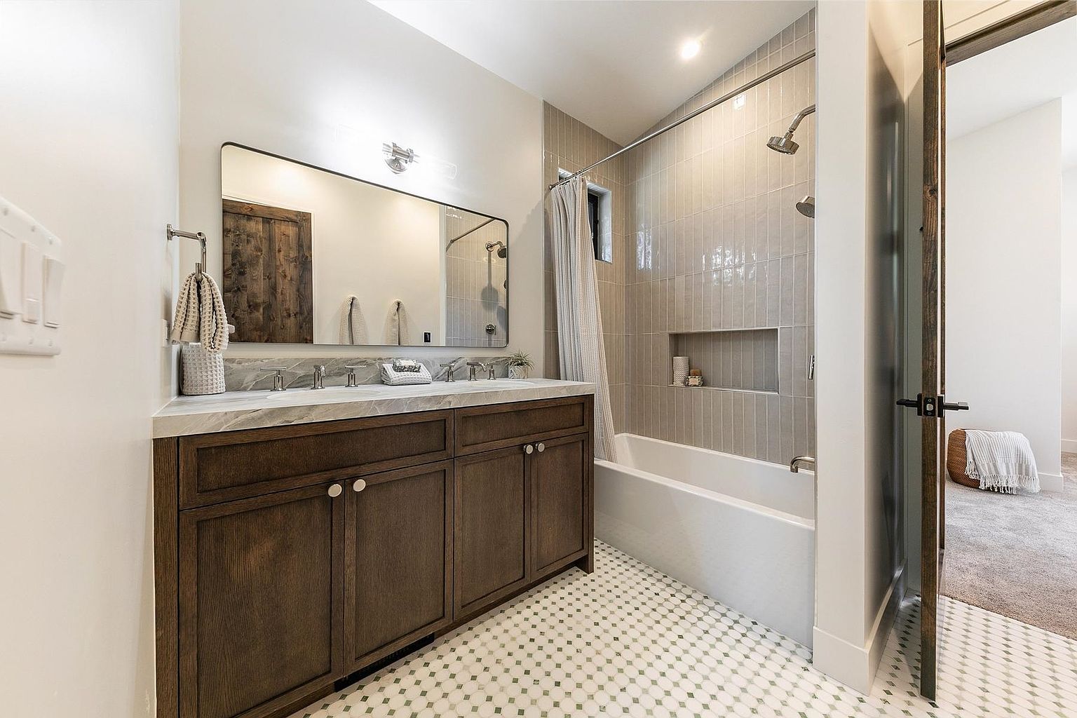 This modern bathroom features a dark wood vanity with a light-colored stone countertop and dual sinks, complemented by a large rectangular mirror. The shower area includes a tub with a built-in niche and vertical light-toned wall tiles, while the floor is finished with a classic patterned tile. The perspective captures the vanity and shower area, highlighting a clean, contemporary aesthetic with a view into an adjacent carpeted room.