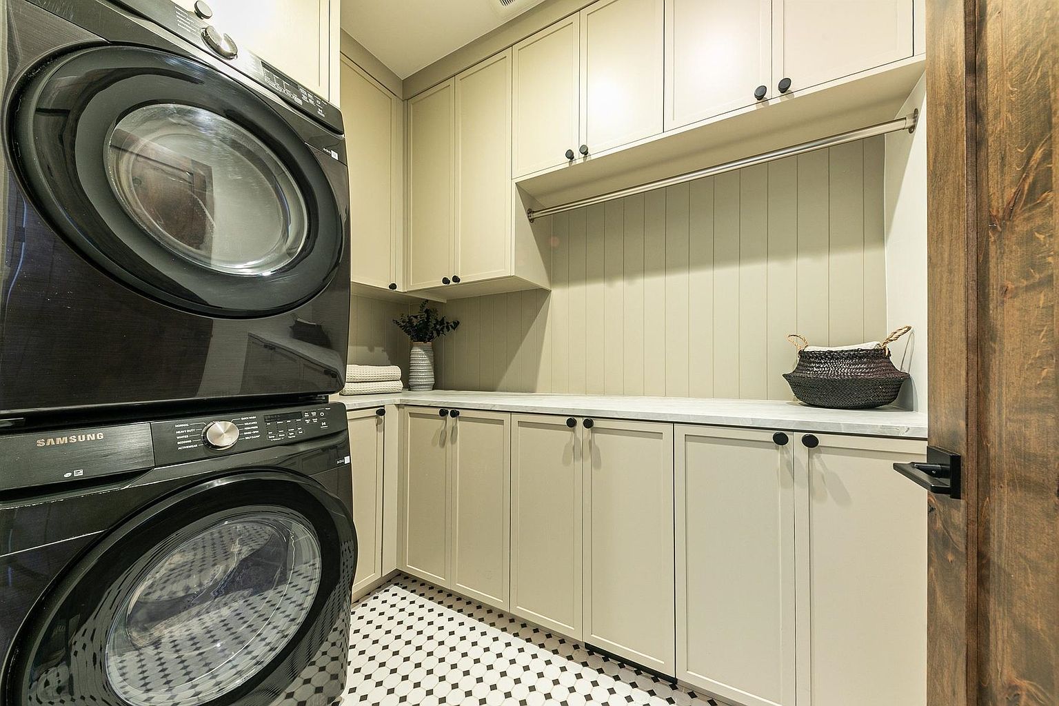 This functional laundry room features a stacked black washer and dryer set positioned next to ample cabinetry with a light-toned finish. A long countertop provides workspace, complemented by a hanging rod and vertical paneling on the wall. The space is finished with a classic black-and-white patterned tile floor, creating a clean and organized aesthetic.