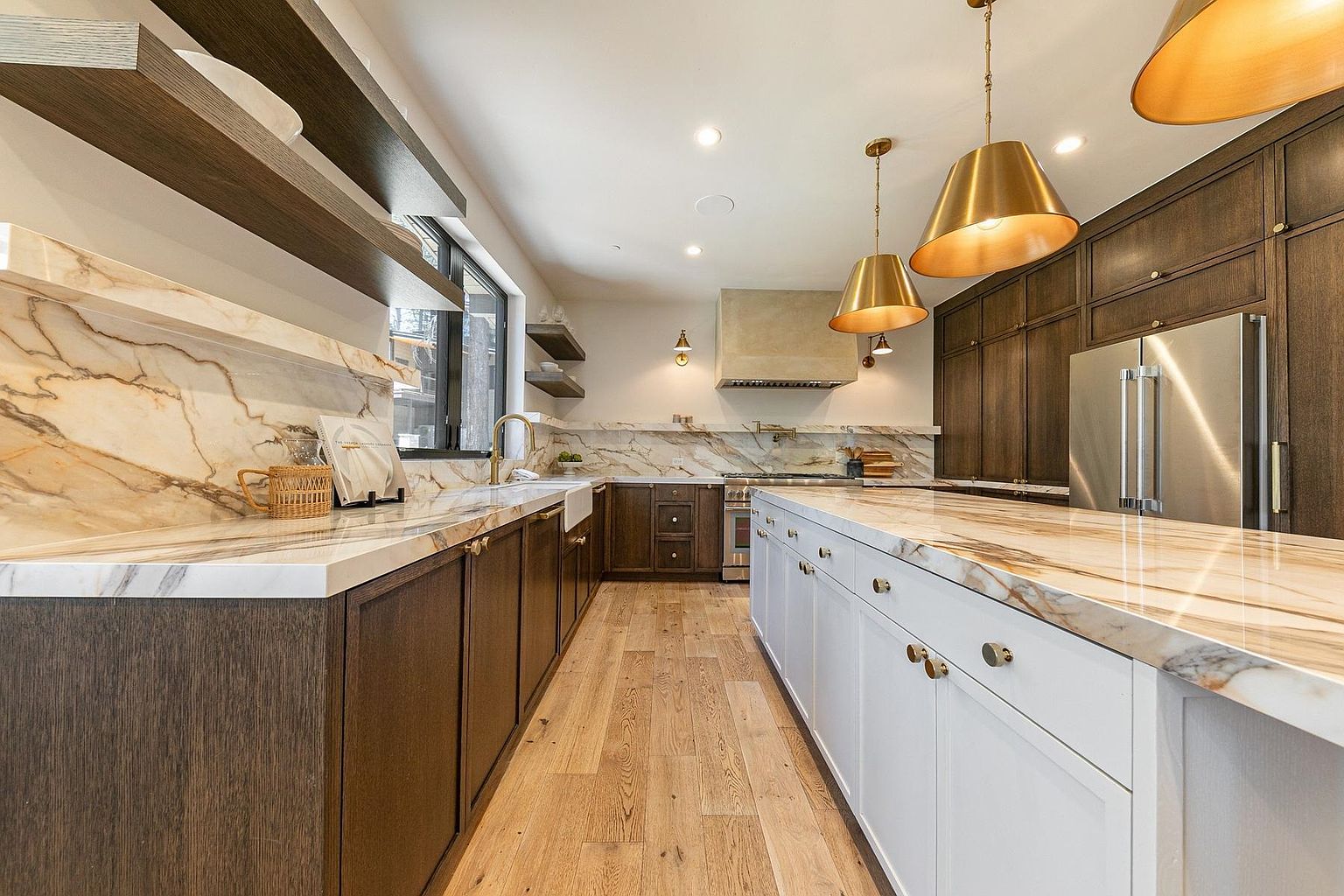 This sophisticated kitchen features a striking contrast between dark wood cabinetry and a bright white kitchen island, both topped with dramatic, veined stone countertops. The space is illuminated by warm, metallic pendant lights hanging over the island, while a custom range hood and open shelving add a refined, modern aesthetic. The perspective is a wide-angle shot from the kitchen entrance, showcasing the seamless flow between the prep areas and the central island.