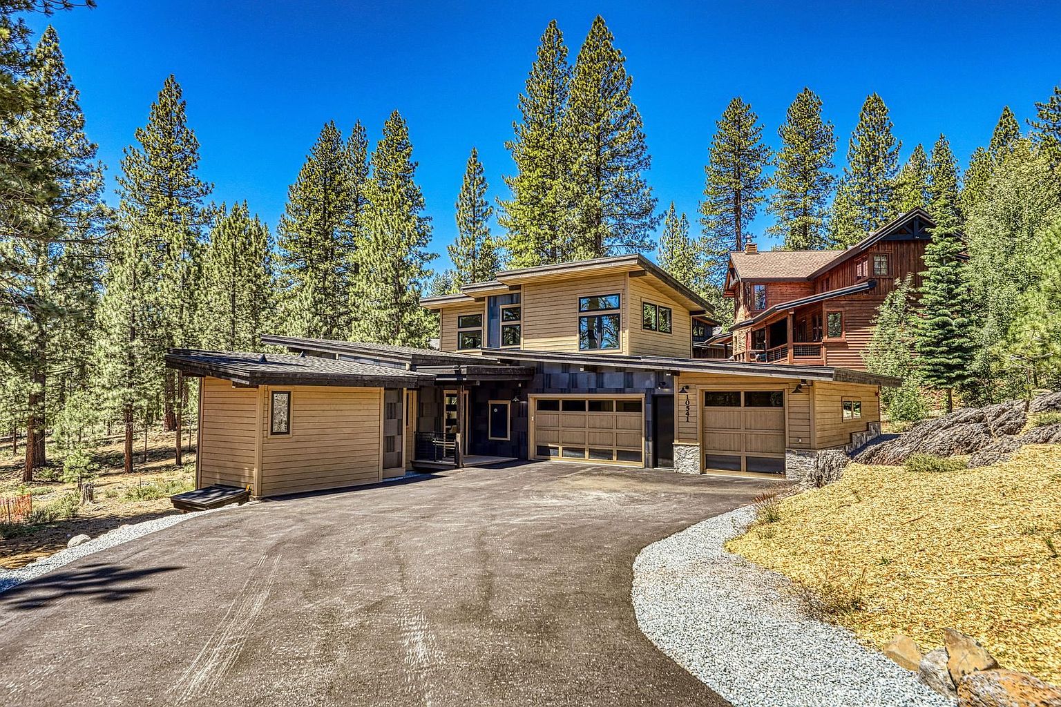 This modern mountain home features a striking blend of horizontal wood siding and dark, textured paneling, set against a backdrop of dense pine trees. The front facade includes a multi-car garage and a recessed entryway, with a paved driveway leading up to the structure. The architectural design emphasizes clean lines and a contemporary aesthetic, perfectly integrated into the natural, wooded landscape.