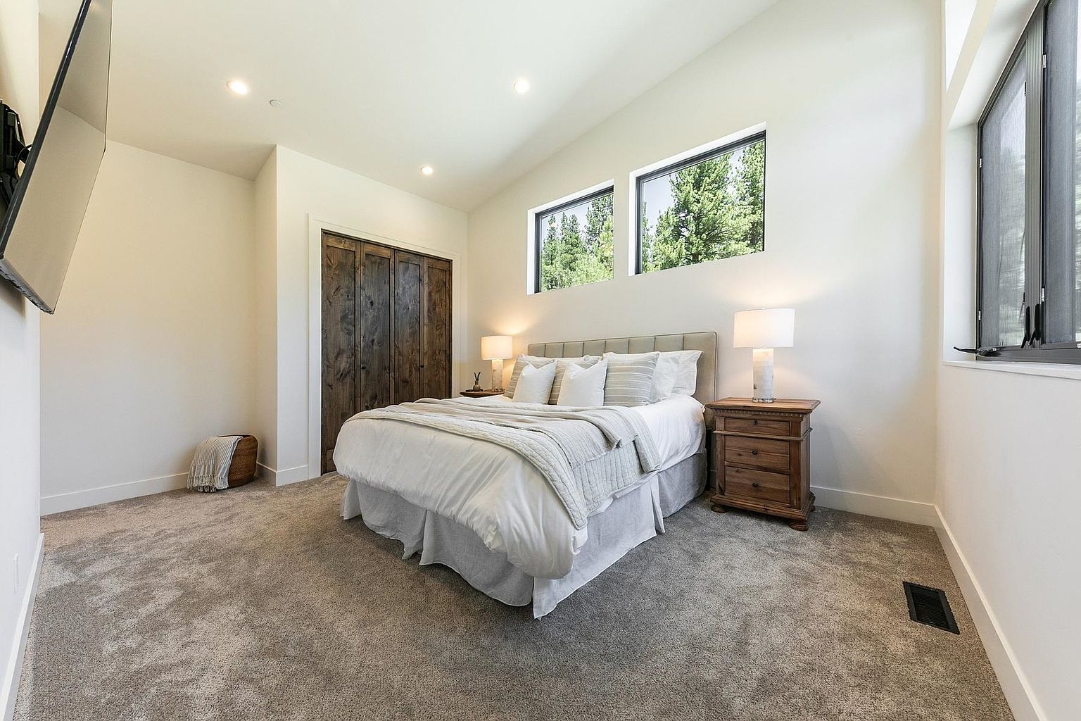 This bright and airy bedroom features a neutral color palette with soft gray carpeting and crisp white walls. A queen-sized bed with a tufted headboard serves as the focal point, flanked by two wooden nightstands with matching table lamps. The room is accented by a rustic dark wood closet door and two rectangular windows that provide natural light and a view of the surrounding trees.