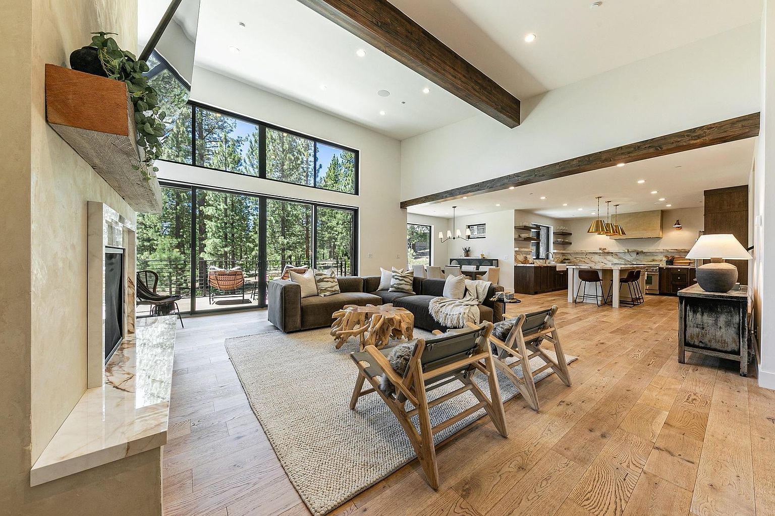 This open-concept living area features a spacious layout with light wood flooring and exposed dark wood ceiling beams. A large, comfortable sectional sofa faces a stone-clad fireplace surround, while a pair of wooden armchairs provides additional seating around a unique organic-shaped coffee table. The space flows seamlessly into a bright kitchen area, with large windows providing an expansive view of the surrounding forest.