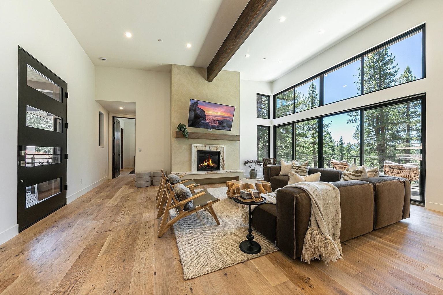 This spacious living room features a modern, open-concept design with light wood flooring and a prominent stone-clad chimney breast centered on the wall. A large sectional sofa and two wooden accent chairs are arranged around a unique, organic-shaped coffee table, creating a cozy seating area. Expansive windows provide abundant natural light and scenic views of the surrounding forest, while a dark-framed glass door adds a striking architectural contrast to the bright, neutral interior.