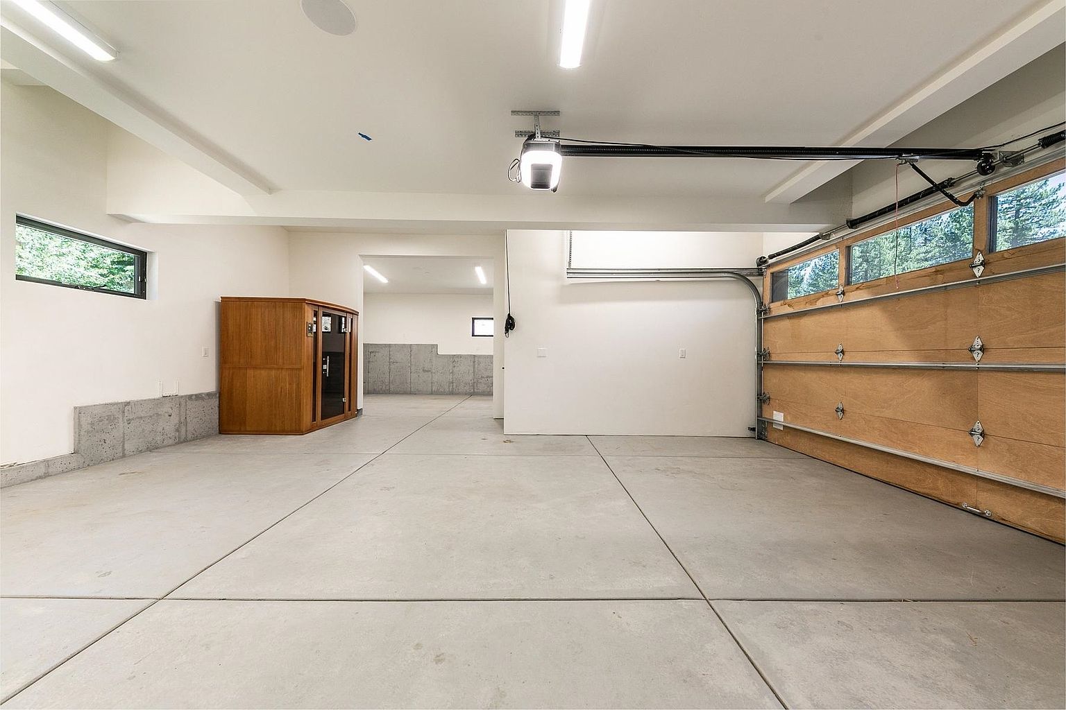 This spacious, well-lit garage features a clean concrete floor and white walls, providing a versatile area for parking or storage. A large wooden garage door with glass panels occupies the right side, while a wooden sauna unit sits against the left wall. The space extends into a secondary, deeper area with concrete foundation walls, offering additional utility or workshop potential.