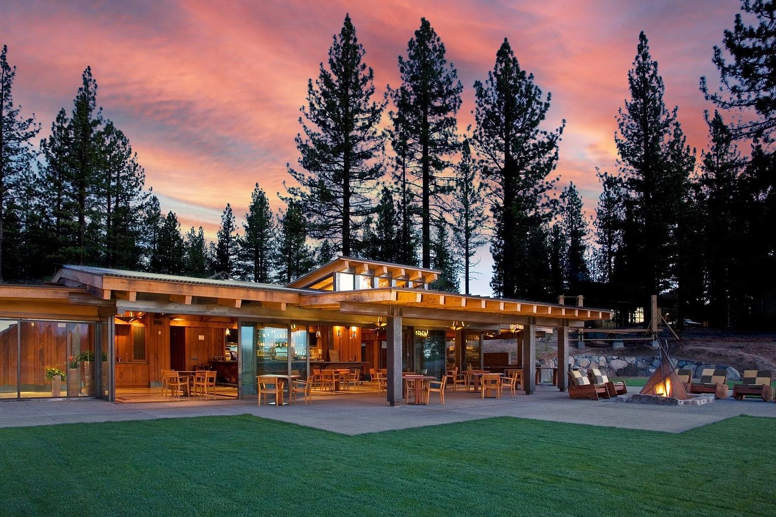 This image captures a modern, wood-clad clubhouse or lodge at dusk, featuring an expansive covered patio area with outdoor dining tables and a central fire pit. The structure utilizes large glass sliding doors that open to the interior, creating a seamless transition between the indoor bar area and the outdoor lawn. The scene is set against a backdrop of tall pine trees under a vibrant, colorful sunset, offering a serene and inviting atmosphere.