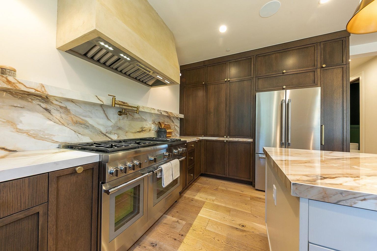 This sophisticated kitchen features dark wood cabinetry paired with a striking stone backsplash and matching countertops that extend to the island. A professional-grade stainless steel range sits beneath a custom-finished range hood, while light-toned wood flooring adds warmth to the space. The perspective captures the functional layout, highlighting the seamless integration of high-end appliances and elegant finishes.